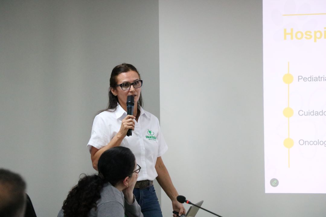 Mulher branca de fios lisos e escuros falando ao microfone na sessão do CFMV. Ela veste calça jeans azul escura e blusa branca de botões no meio com nome da Inataa.