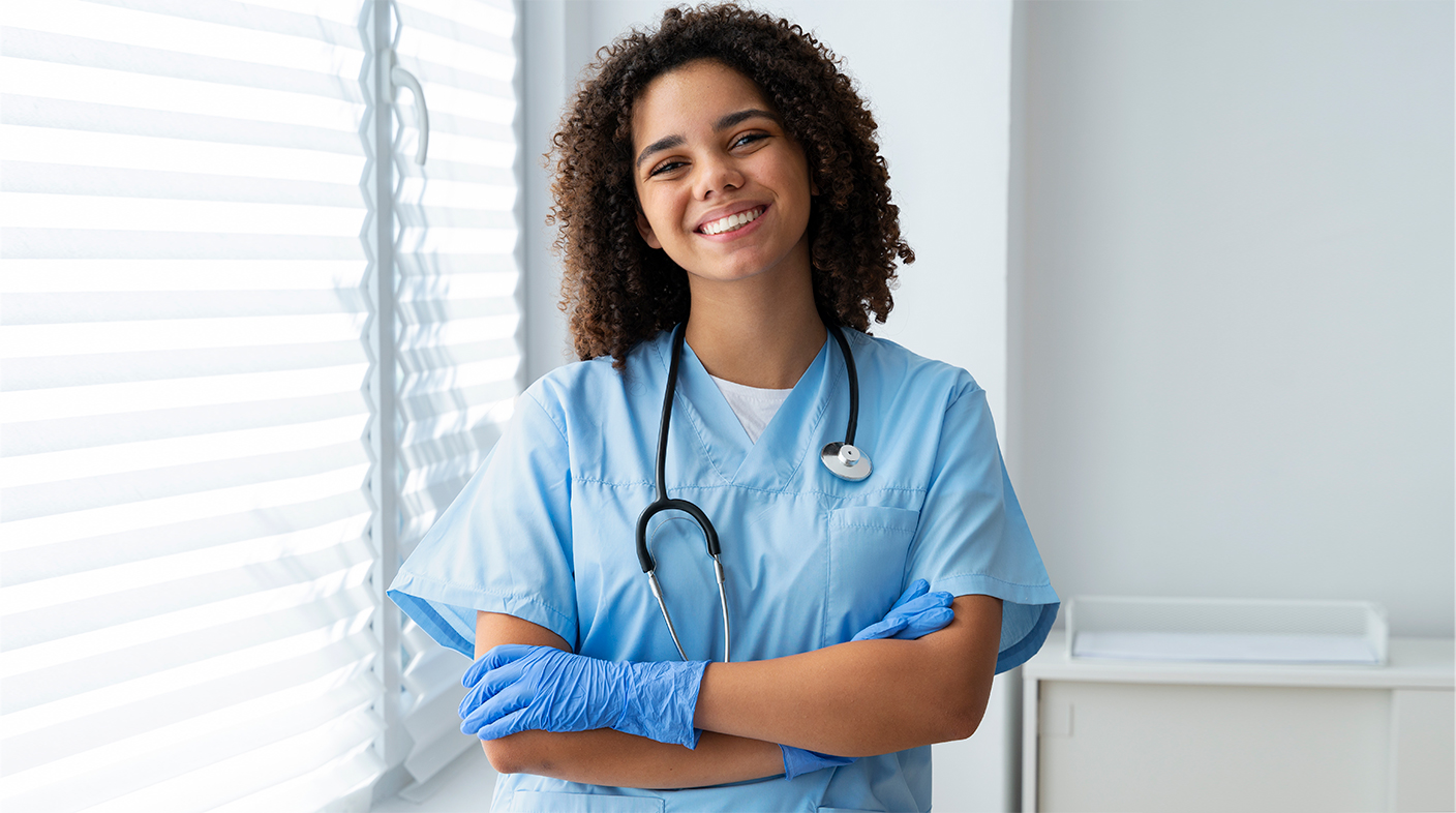 Mulher jovem, negra de cabelos cacheados na altura dos ombros, sorrindo para foto. Ela veste uniforme e luvas cirúrgicas azuis e tem um estetoscópio no pescoço.