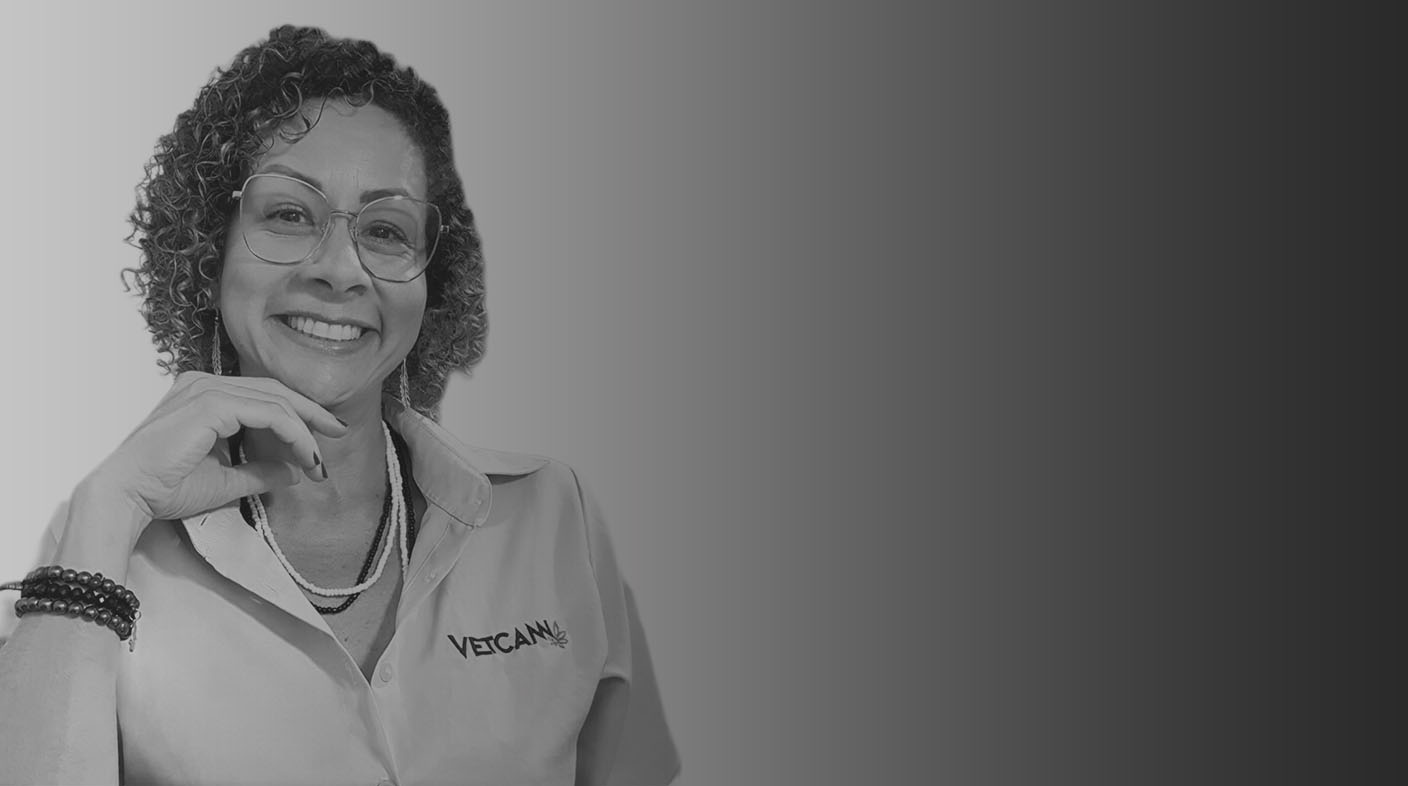 Mulher de cabelos cacheados curtos usando óculos de grau sorri para foto. Ela veste jaleco branco de médico-veterinário, usa relógio na mão esquerda e está com a mão esquerda abaixo do queixo. É a dra. Aline Goulart.