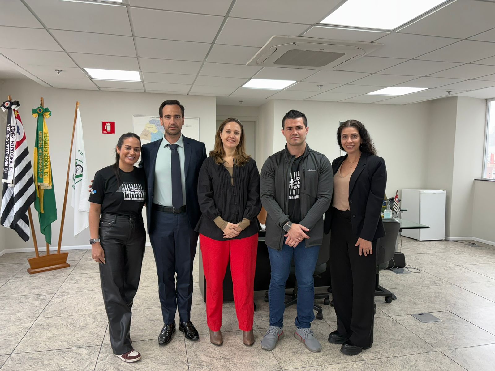 Membros da Diretoria pousando para foto com deputados da causa animal. Cinco pessoas de pé, sendo três mulheres e dois homens, em uma sala de reunião, sorrindo em sala de reunião com as bandeiras do estado de São Paulo, do Brasil e do CRMV-SP ao fundo, à esquerda.