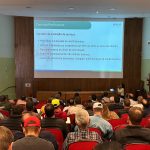 Visão das costas do público sentado em cadeiras vermelhas em um auditório olhando o telão.