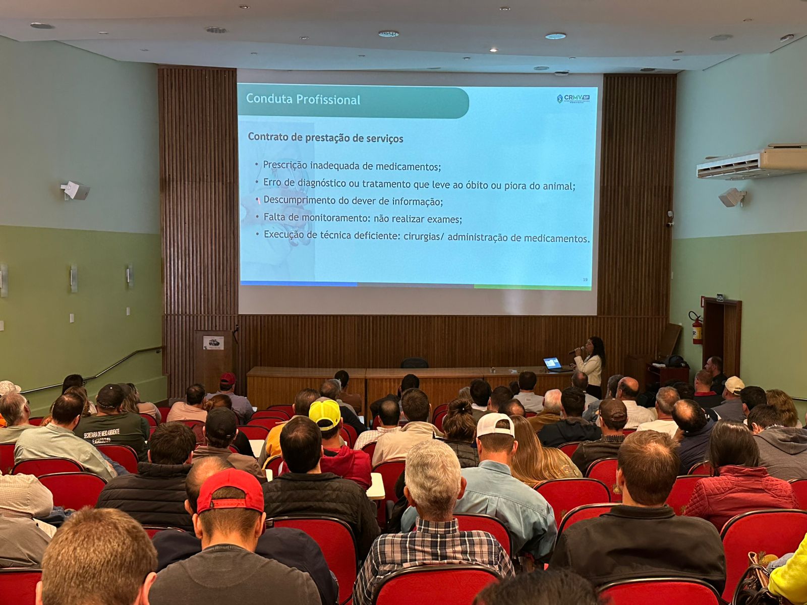 Visão das costas do público sentado em cadeiras vermelhas em um auditório olhando o telão.