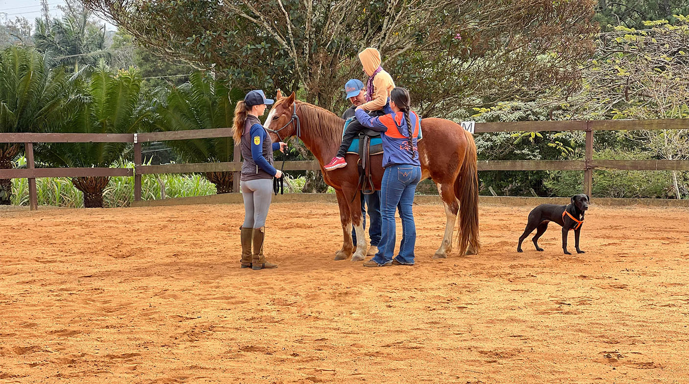 Um paciente de Equoterapia, sobre um cavalo, com três profissionais o auxiliando, em um chão de terra, com uma cerca e árvores ao fundo e um cachorro próximo ao cavalo.