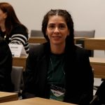 Mulher branca de cabelos longos, cacheados e pretos, sentada atrás de uma mesa sorrindo para foto. É a vice-presidente do CRMV-SP, Carolina Filippos.