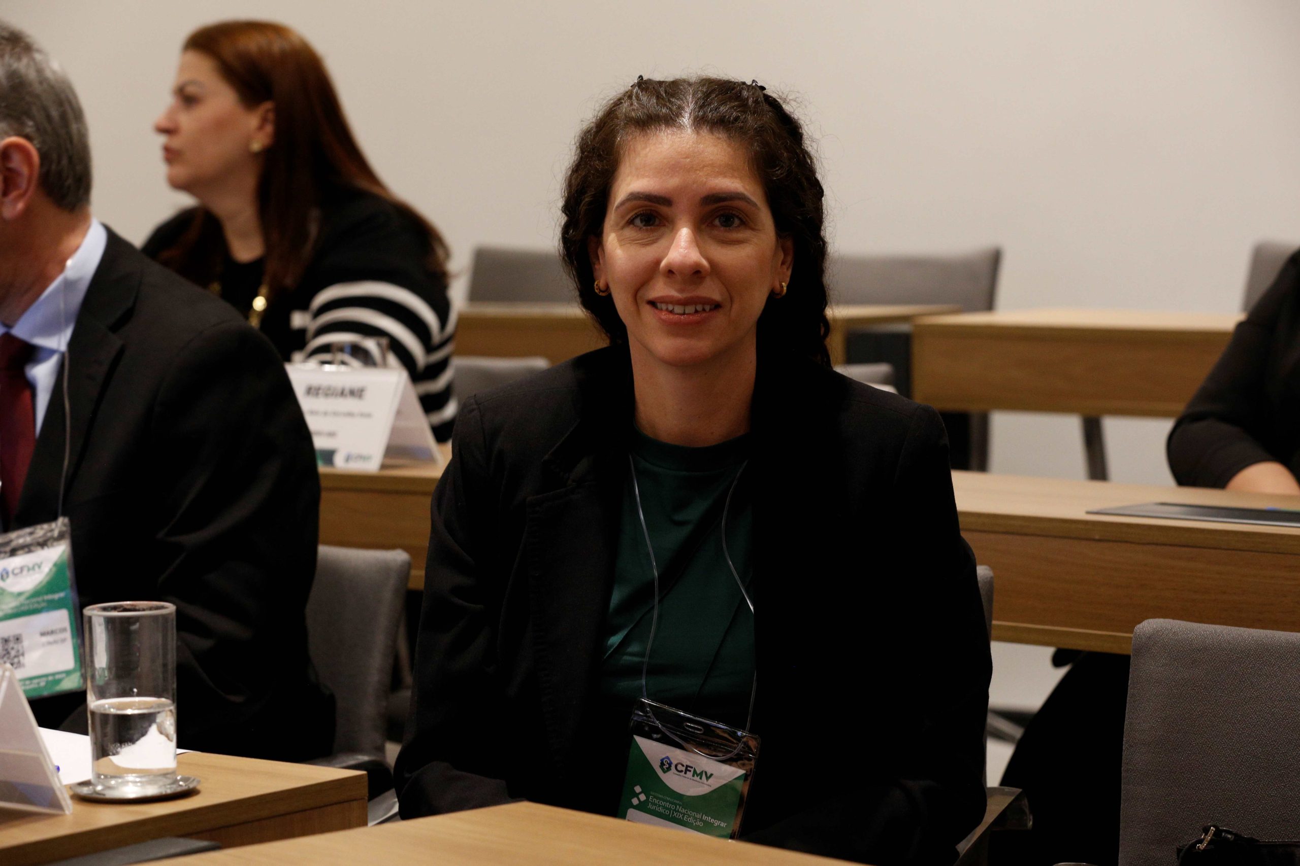 Mulher branca de cabelos longos, cacheados e pretos, sentada atrás de uma mesa sorrindo para foto. É a vice-presidente do CRMV-SP, Carolina Filippos.