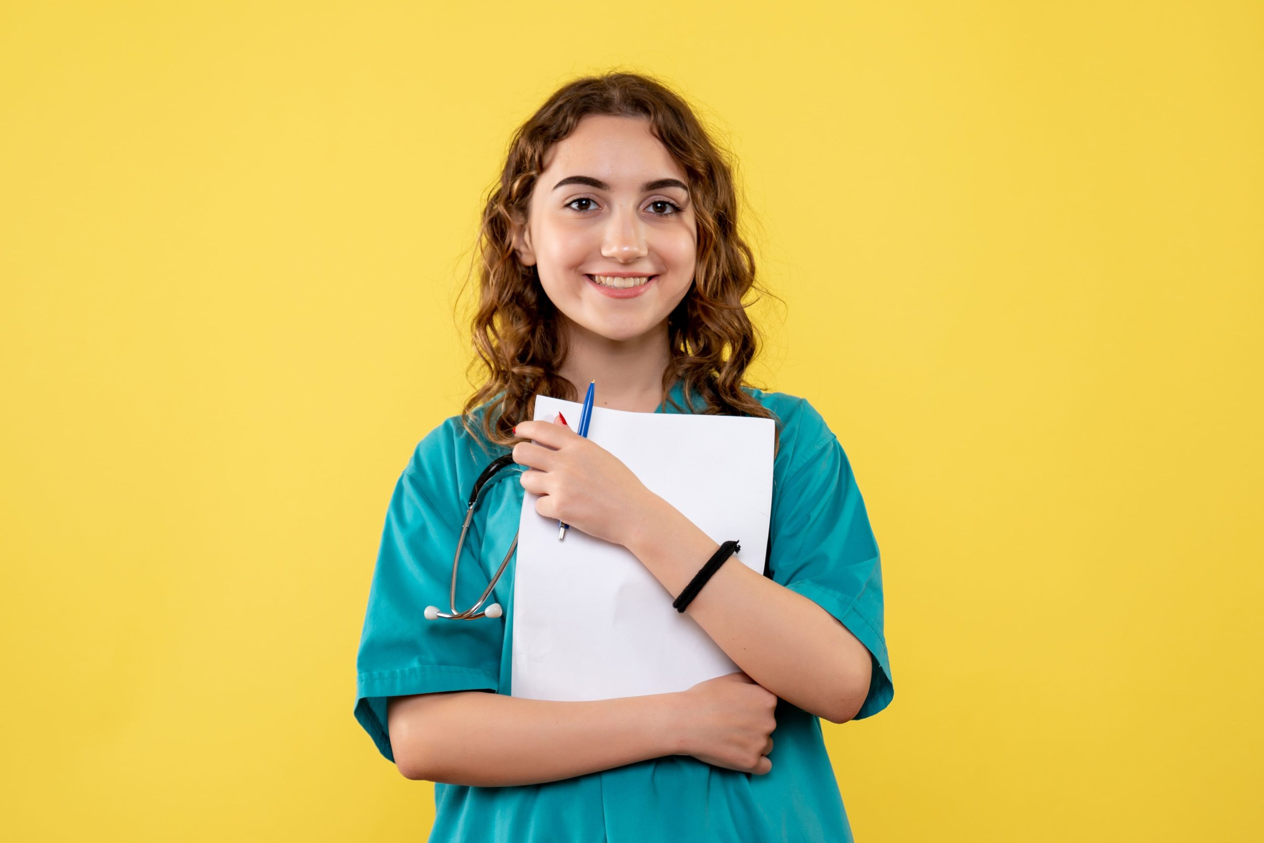 Mulher jovem branca de fios ondulados loiro escuro sorri enquanto segura papéis e uma caneta enquanto veste uniforme verde de médico-veterinário e estetoscópio no pescoço.