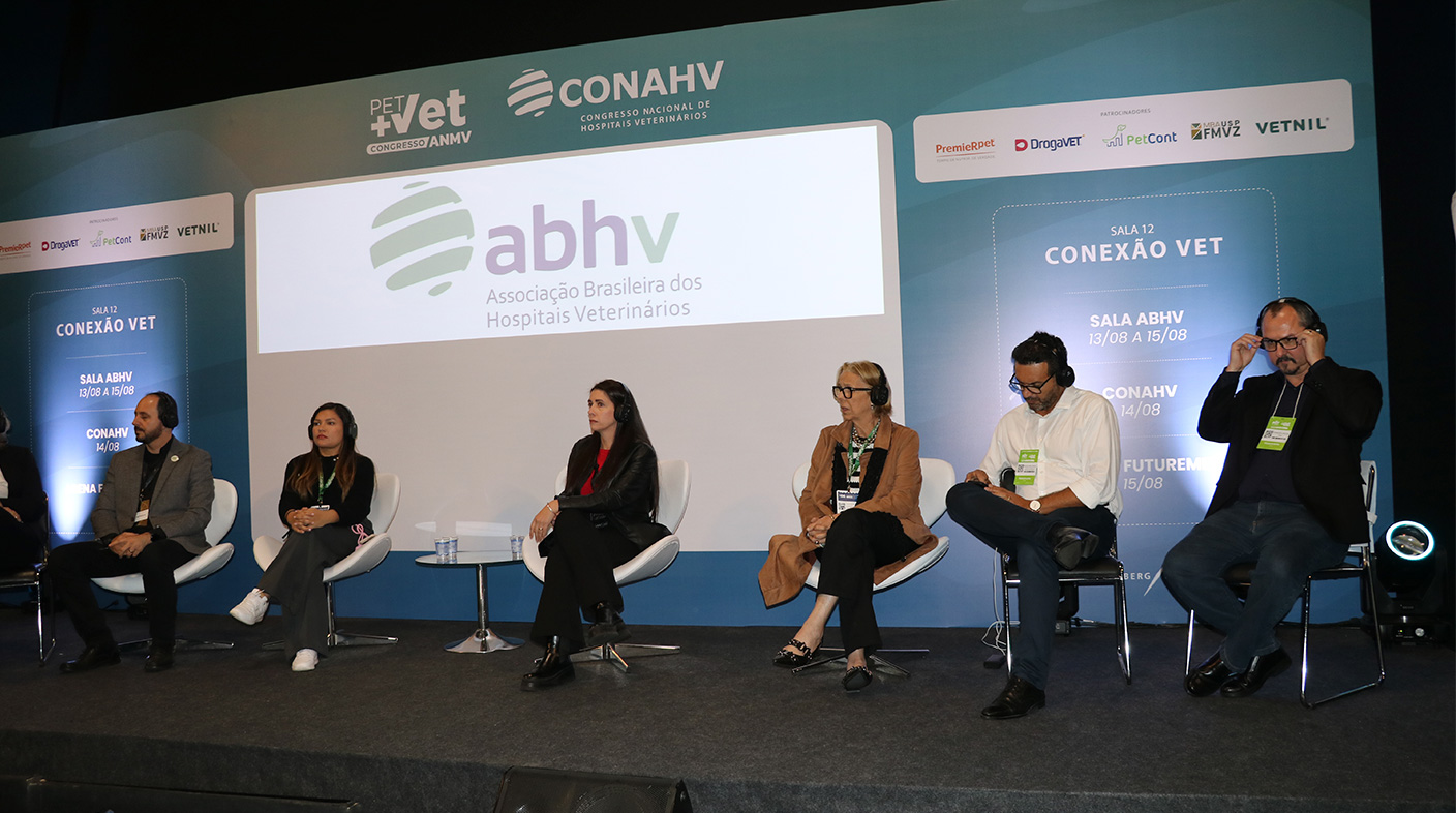 Seis pessoas sentadas lado a lado, em cima de um palco, cada uma em uma cadeira branca atrás delas, o logo da ABHV(Associação Brasileira dos Hospitais Veterinários).