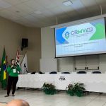 Mulher falando frente a mesa e telão.