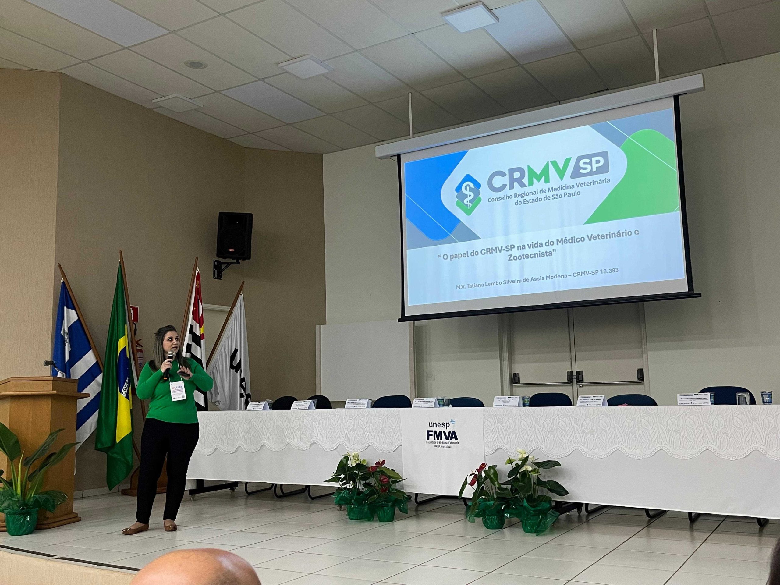 Mulher falando frente a mesa e telão.