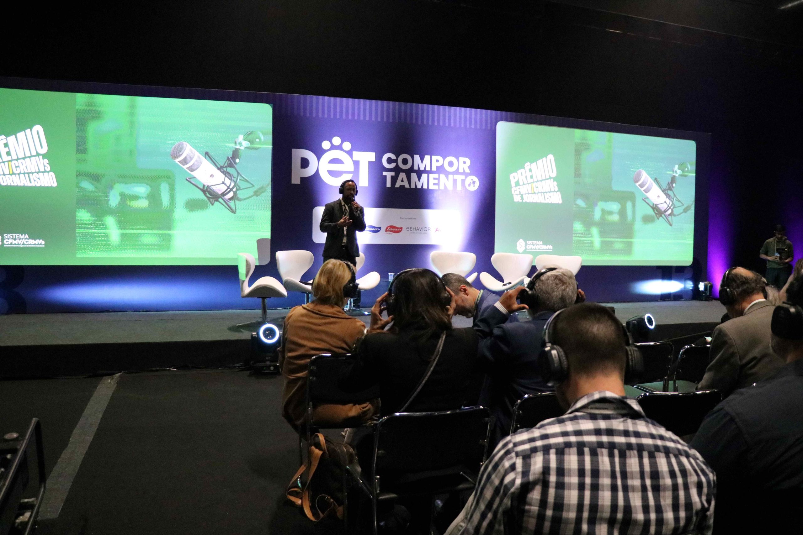 Homem em pé em cima de um palco, falando em microfone para plateia.
