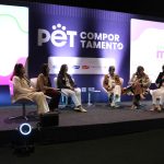 Seis mulheres sentadas em cadeiras no palco a frente do telão.