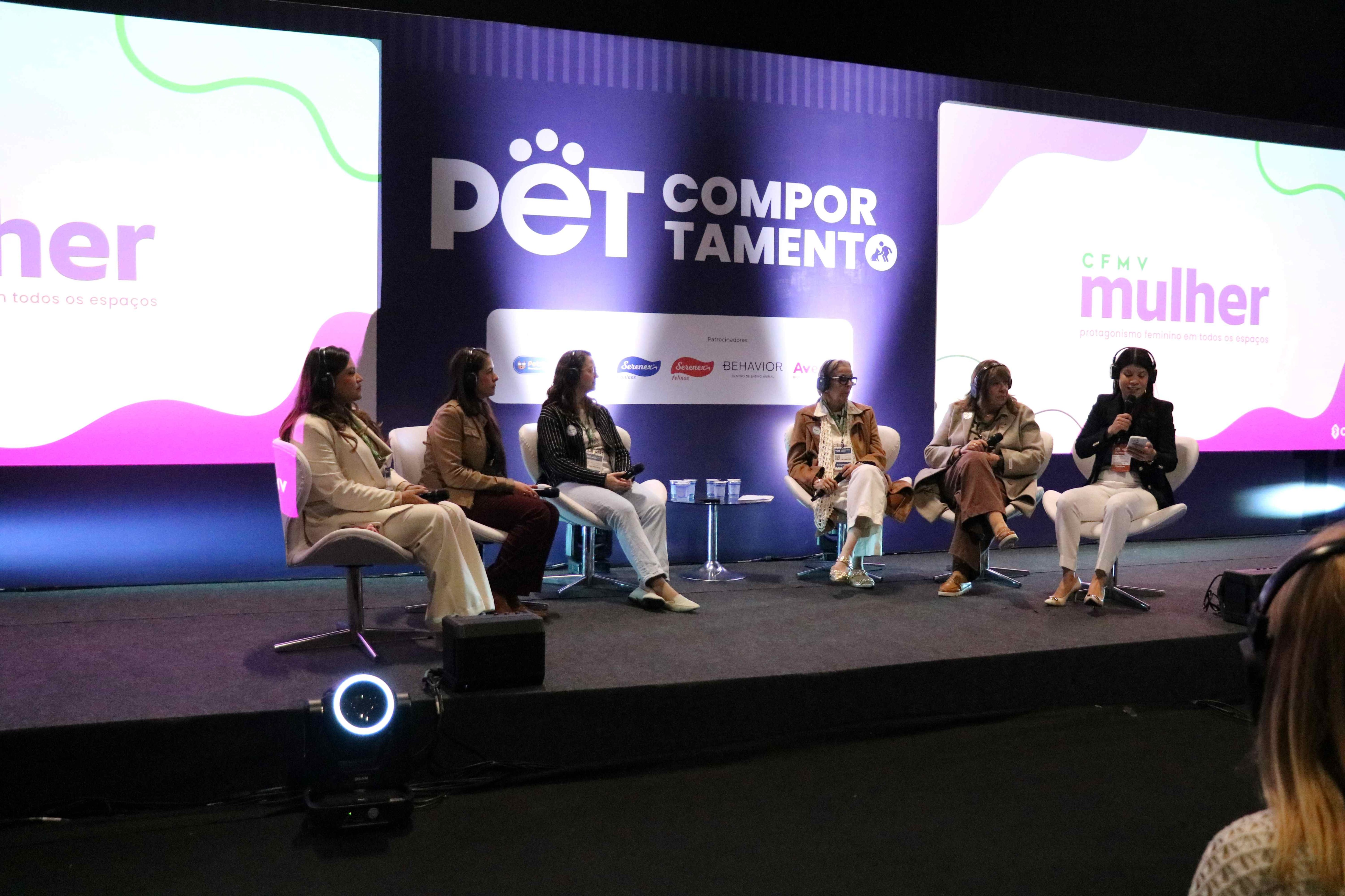 Seis mulheres sentadas em cadeiras no palco a frente do telão.