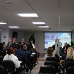 Visão das costas do público com o braço levantado interagindo com a palestrante.