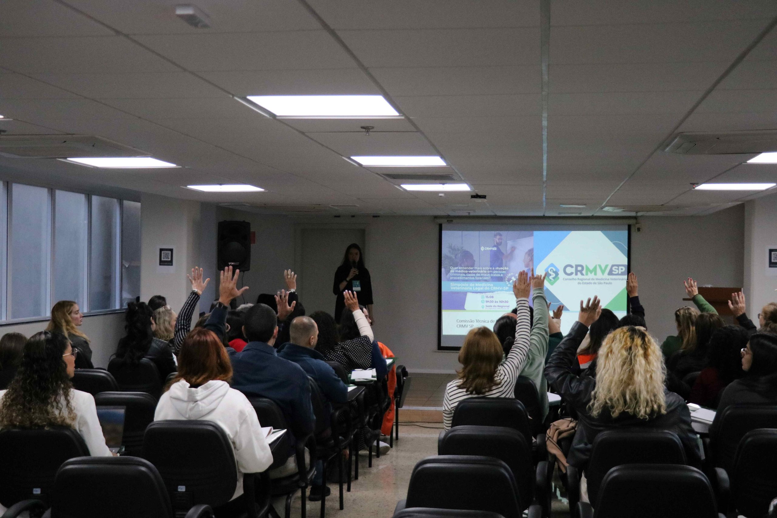 Visão das costas do público com o braço levantado interagindo com a palestrante.
