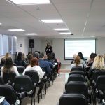 Visão das costas do público olhando mulher em frente ao telão falando no microfone para público sentado.