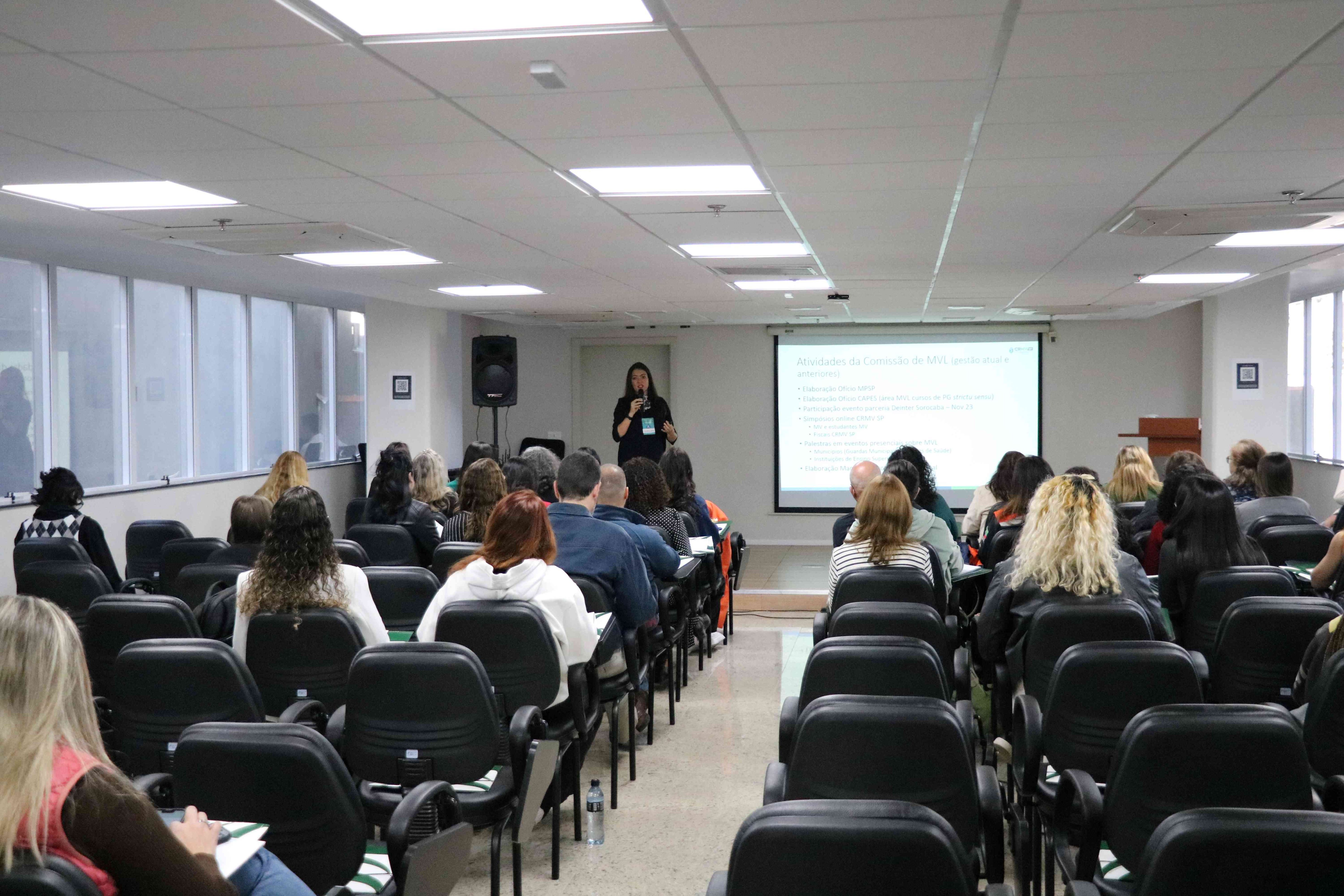Visão das costas do público olhando mulher em frente ao telão falando no microfone para público sentado.