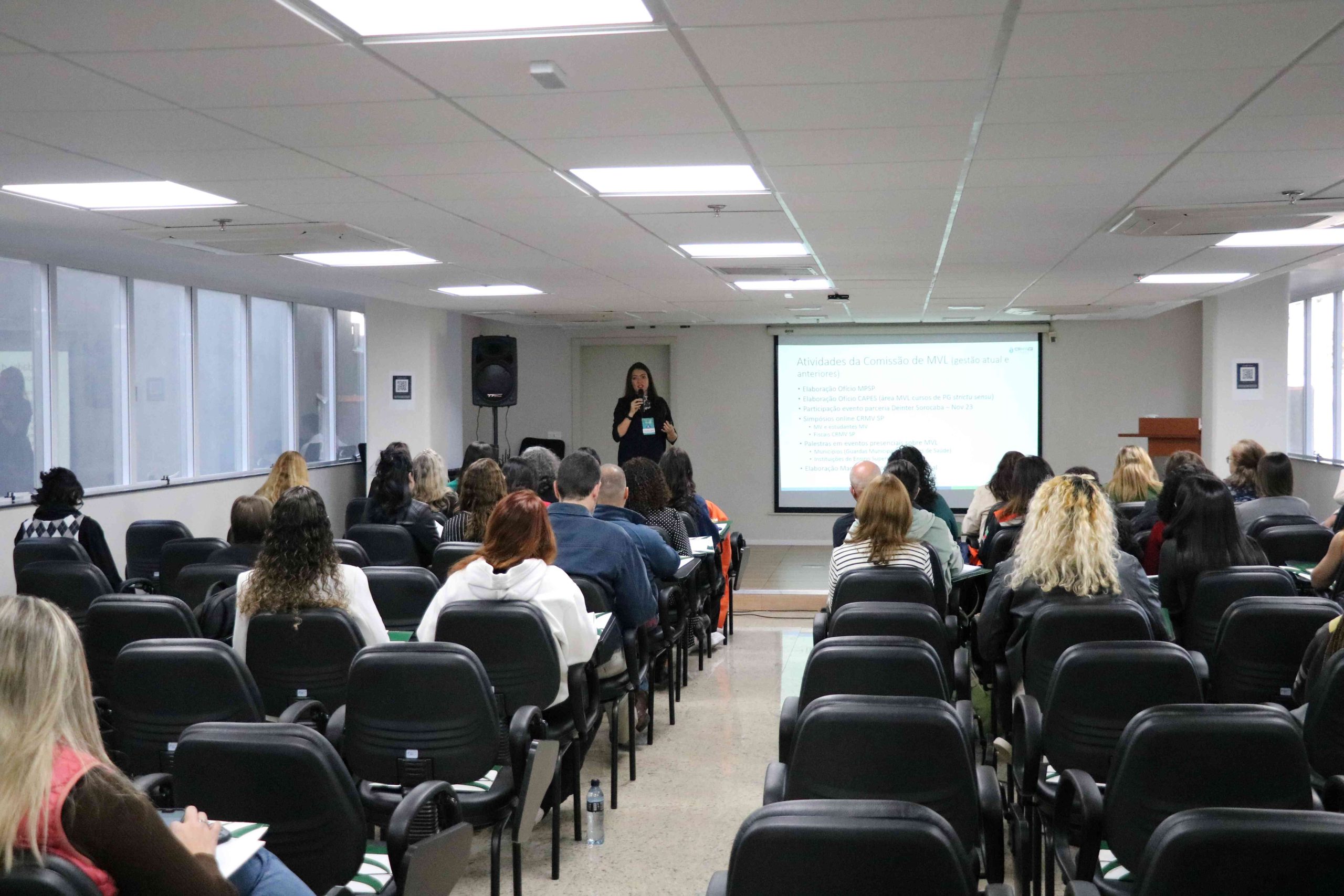 Visão das costas do público olhando mulher em frente ao telão falando no microfone para público sentado.