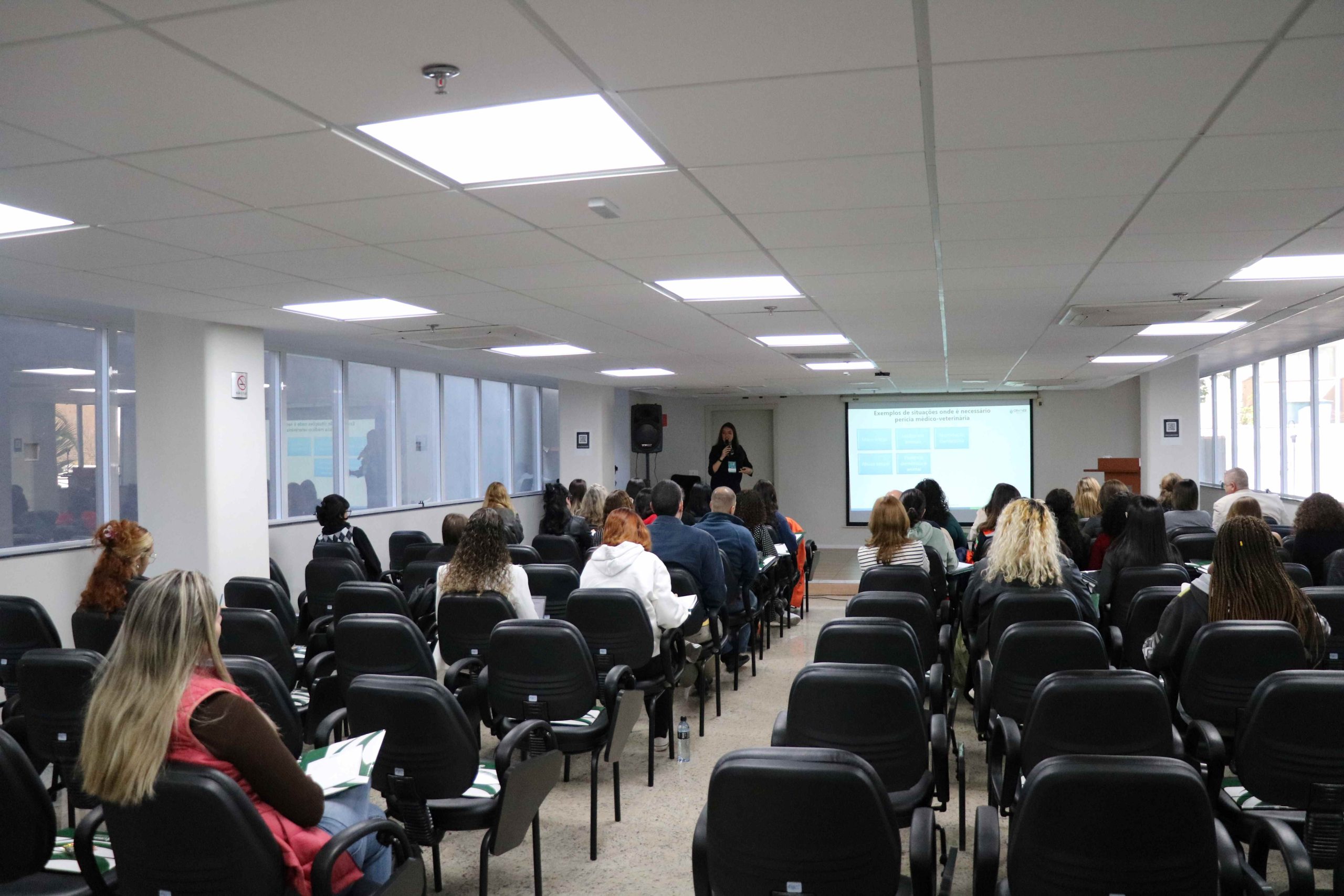 Visão das costas do público sentado assistindo palestra ministrada por mulher.