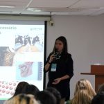 Mulher ao lado de telão falando ao microfone para público.