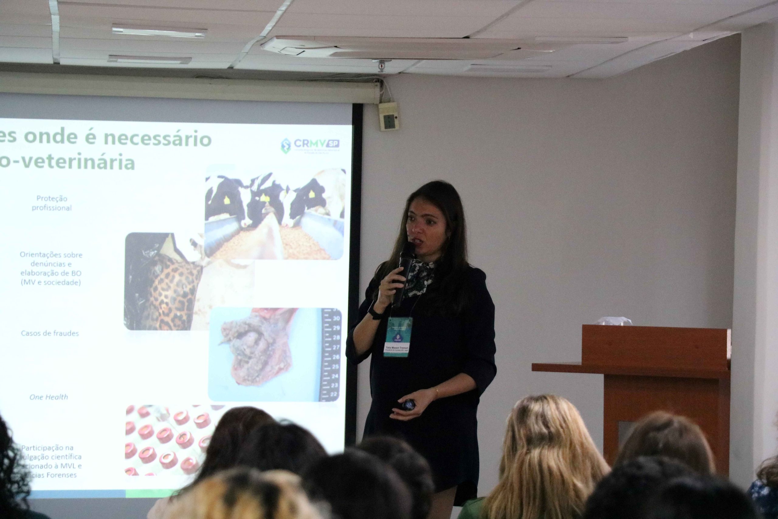 Mulher ao lado de telão falando ao microfone para público.