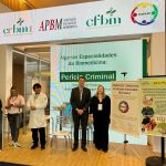 Homem e mulher em pé posando para foto em fachada de evento.