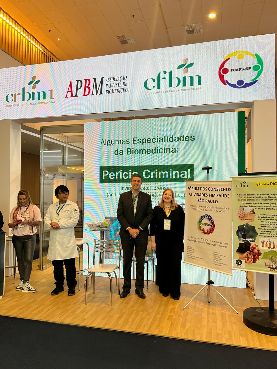 Homem e mulher em pé posando para foto em fachada de evento.