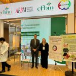 Homem e mulher em pé posando para foto em fachada de evento.