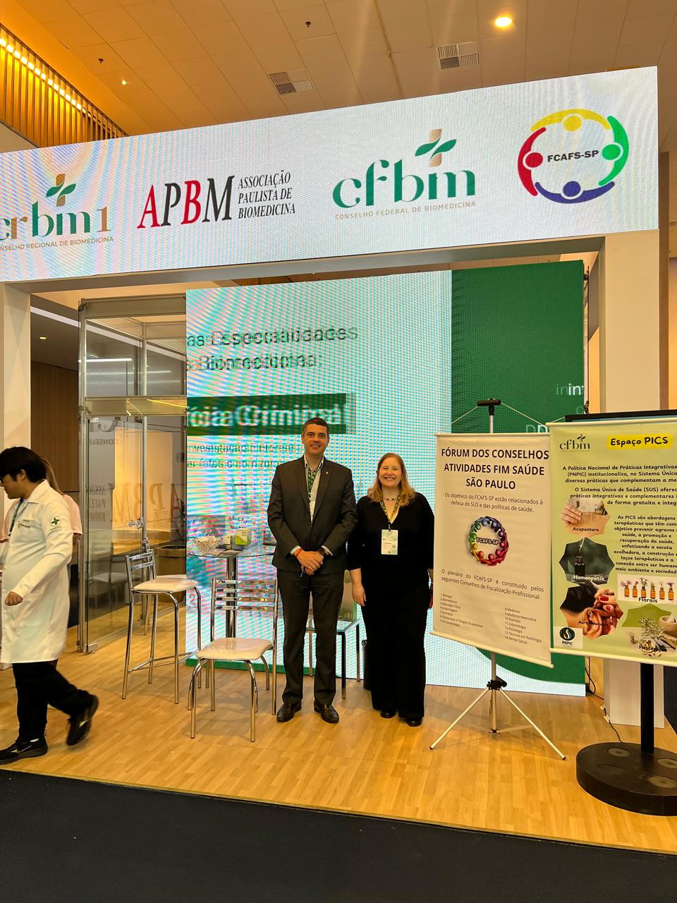 Homem e mulher em pé posando para foto em fachada de evento.