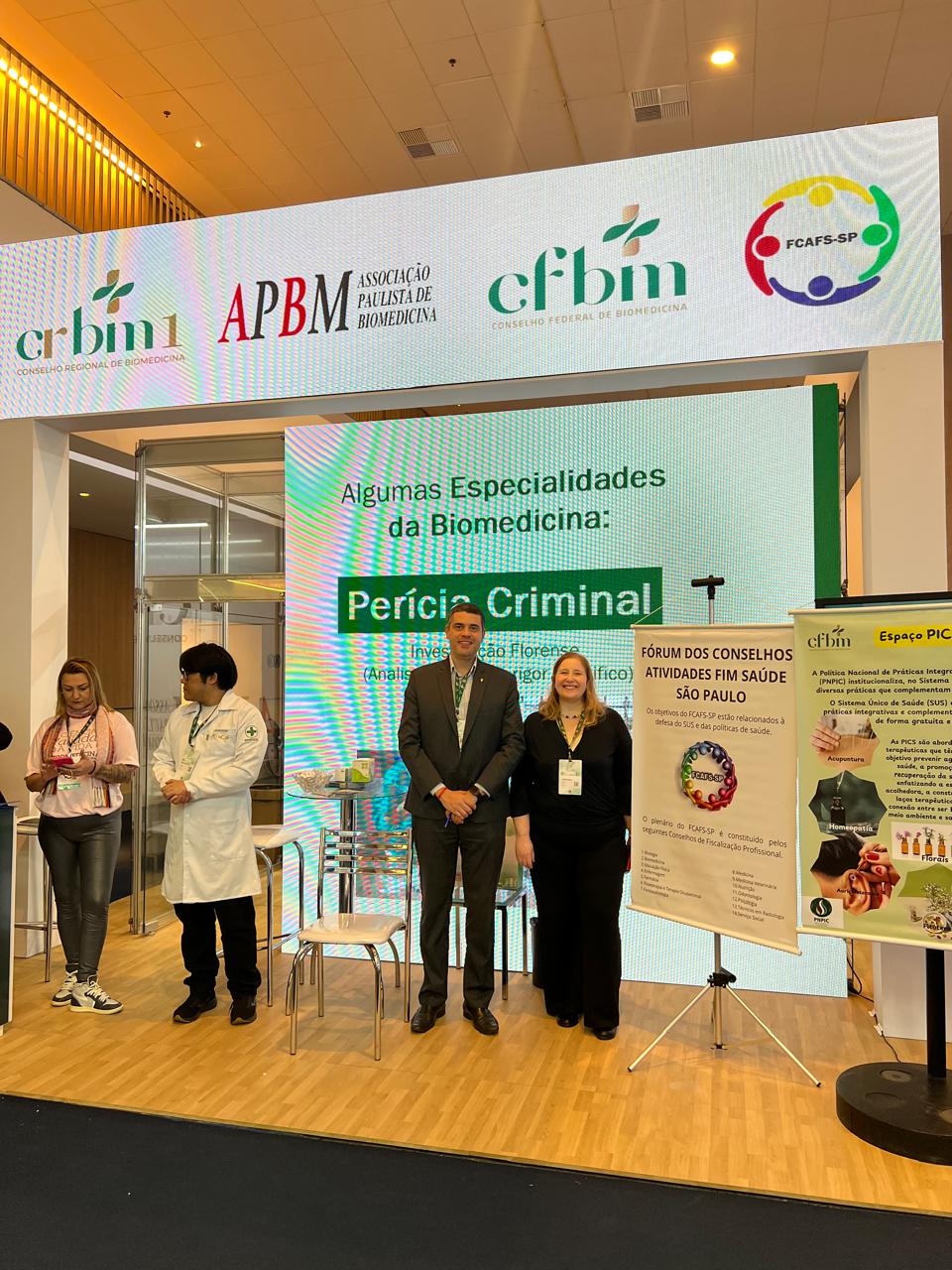 Homem e mulher em pé posando para foto em fachada de evento.