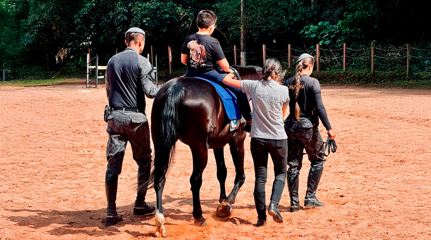 Um paciente de equoterapia sobre um cavalo, de costas, com três profissionais acompanhando a marcha do animal, em um espaço de terra, ao fundo uma cerca e árvores.