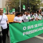 Manifestantes vestindo blusa branca segurando cartazes e uma faixa retangular verde com os dizeres em letras brancas: O SUS se faz com presencialidade.