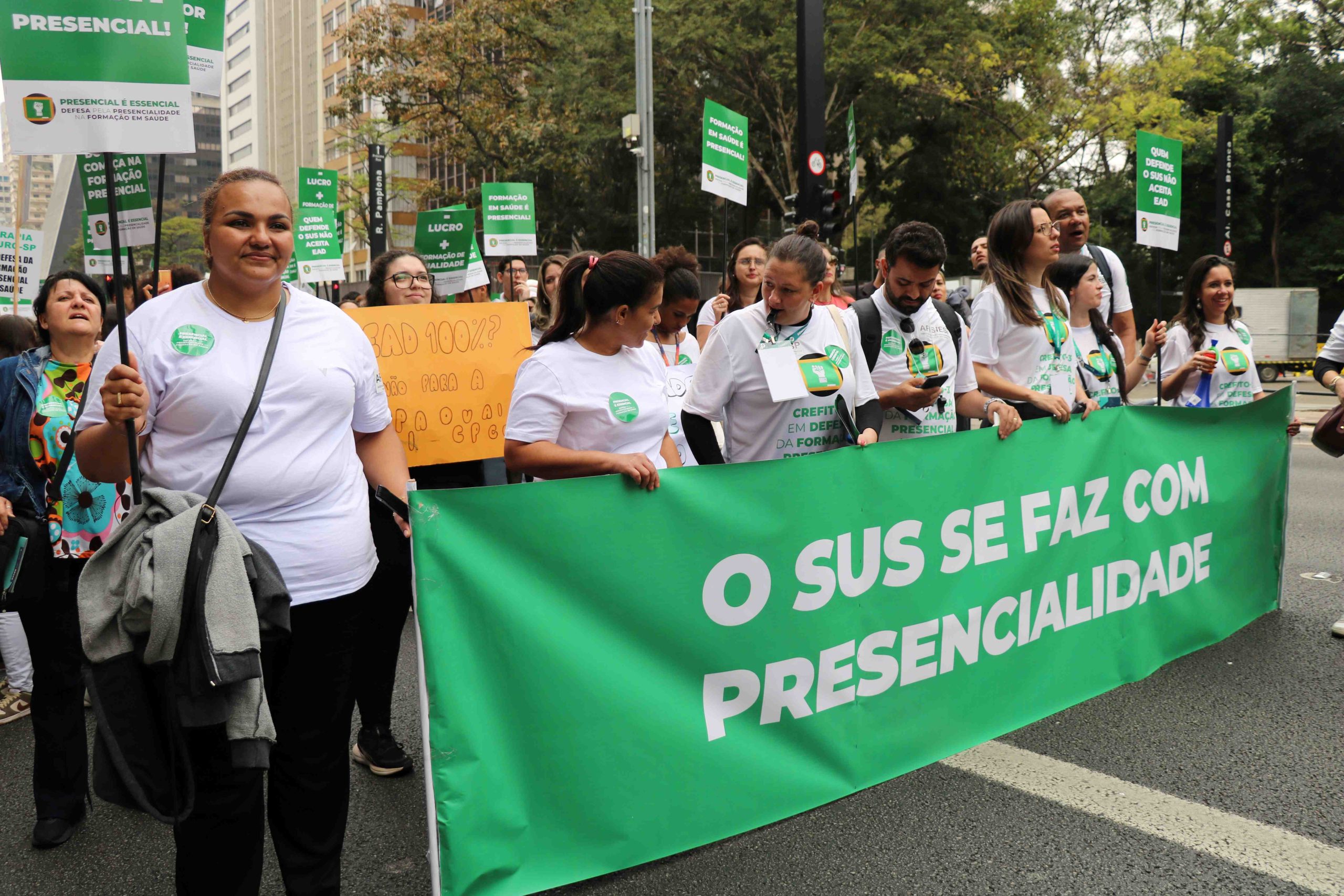 Manifestantes vestindo blusa branca segurando cartazes e uma faixa retangular verde com os dizeres em letras brancas: O SUS se faz com presencialidade.