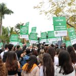 Pessoas com blusas brancas andando em passeata com placas em defesa do ensino presencial na área da Saúde.
