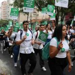 Imagem frontal mostrando estudantes subindo a rua, vestindo blusas brancas, usando apitos e segurando placas com os dizeres: Menos Lucro, Mais formação de Qualidade.