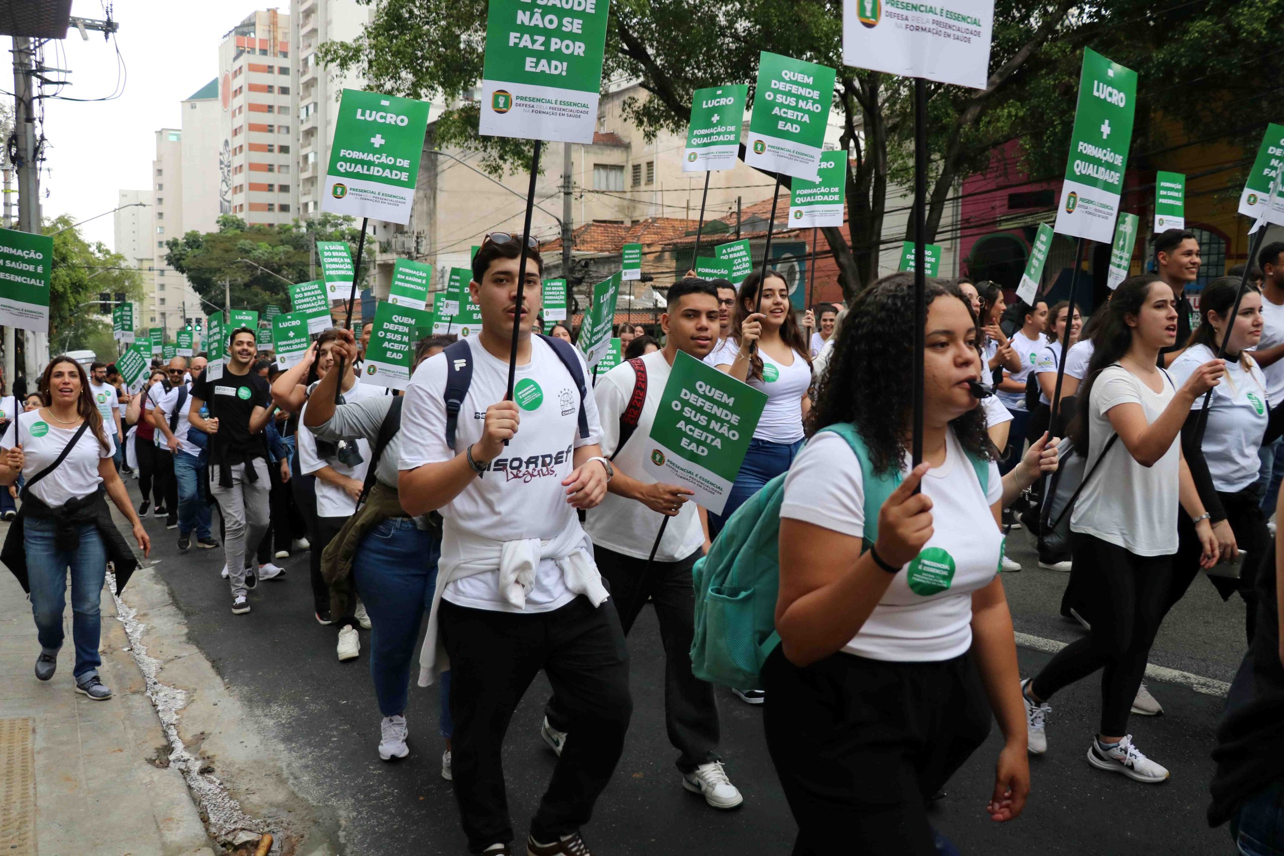 Imagem frontal mostrando estudantes subindo a rua, vestindo blusas brancas, usando apitos e segurando placas com os dizeres: Menos Lucro, Mais formação de Qualidade.