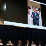 Pesosas sentada atrás de uma mesa retangular em cima de um palco preto, enquato um vídeo é exibido em um telão quadrado acima delas.