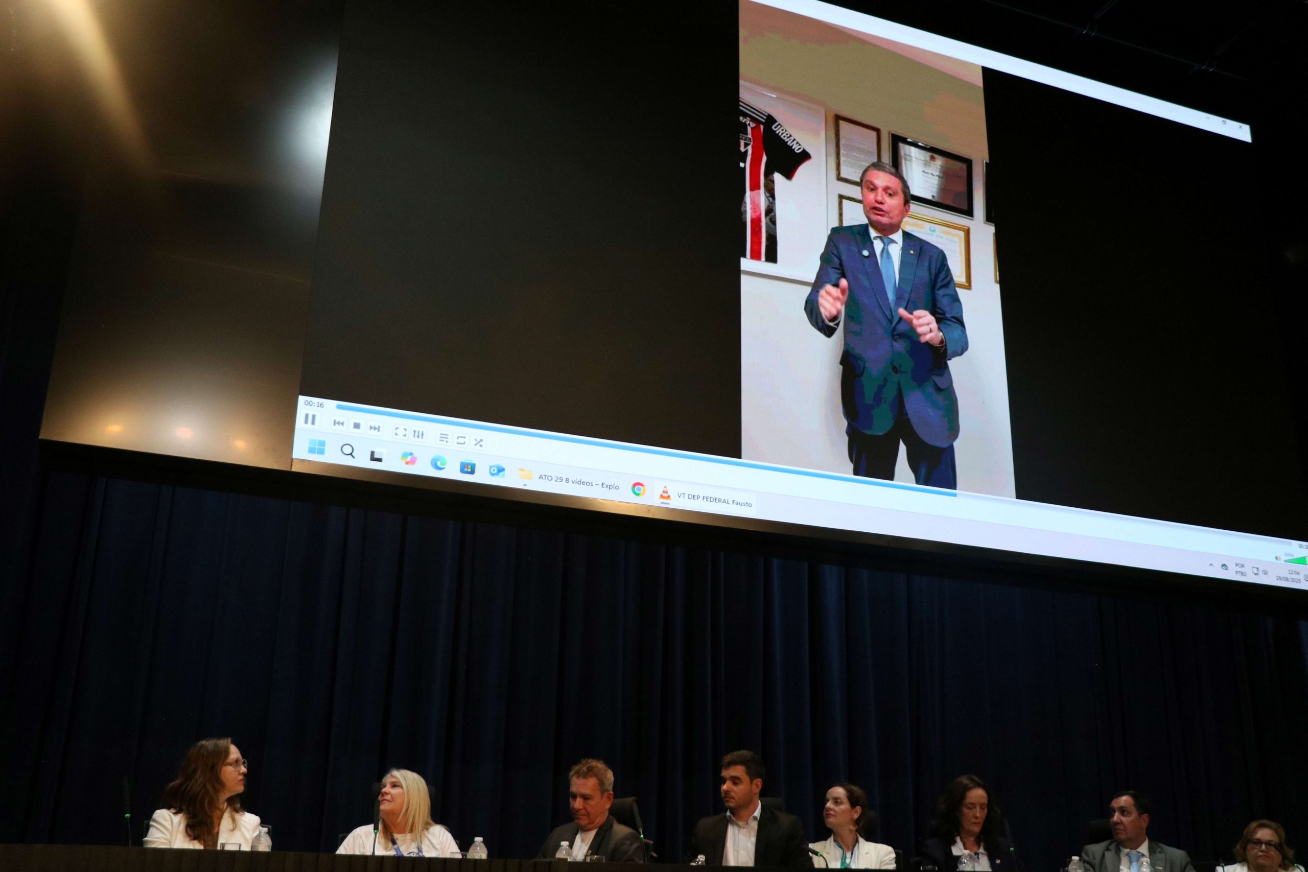 Pesosas sentada atrás de uma mesa retangular em cima de um palco preto, enquato um vídeo é exibido em um telão quadrado acima delas.