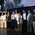Nove autoridades em pé em cima de um palco preto em auditório oficial na Alesp, com uma bandeira do estado de São Paulo ao fundo à esquerda.