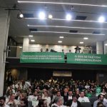 Naimagem, pessoas sentadas na plateia e no mezanino da sala Sandro Montoro na Alesp. No mezanino, faixas verdes com dizeres em letras brancas em favor do SUS.