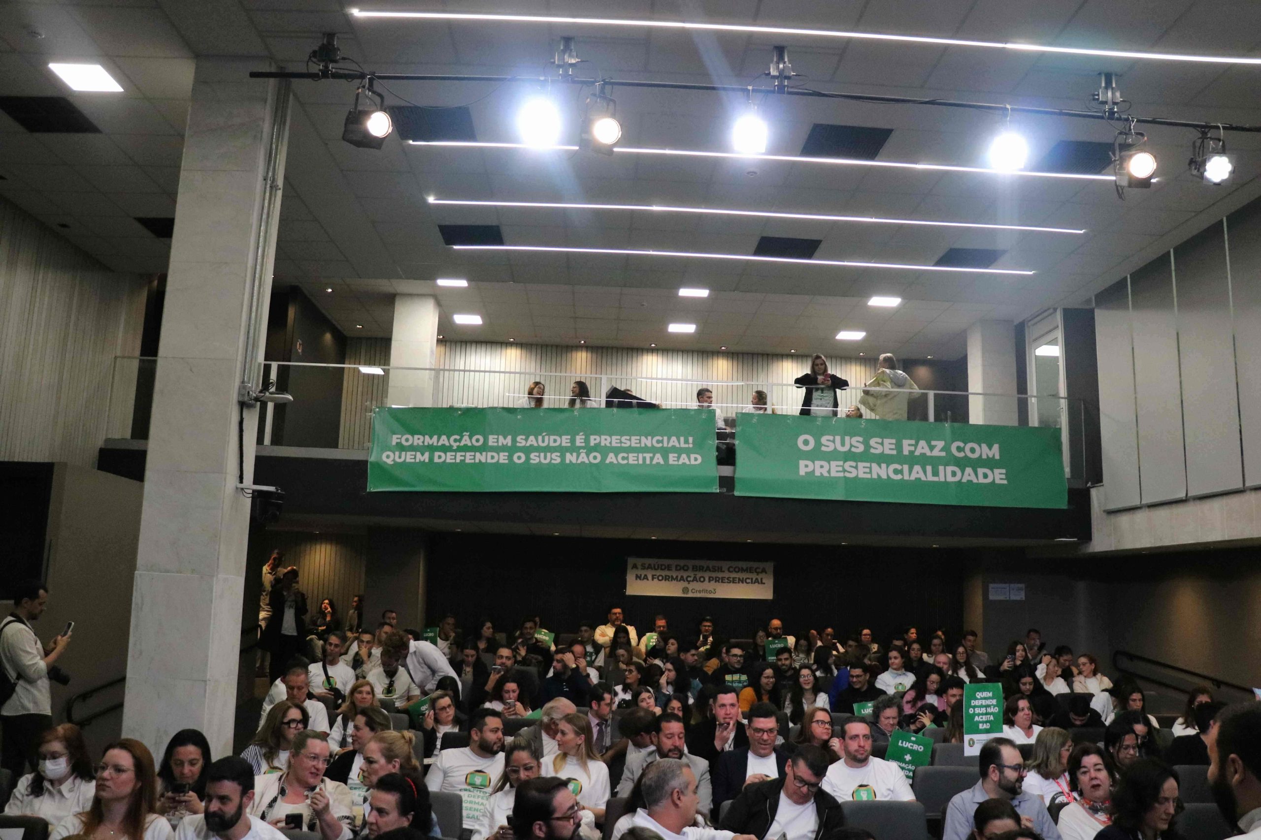Naimagem, pessoas sentadas na plateia e no mezanino da sala Sandro Montoro na Alesp. No mezanino, faixas verdes com dizeres em letras brancas em favor do SUS.