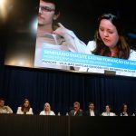 A presidente do CRMV-SP, Daniela Pontes Chiebao, sentada na mesa principal do seminário discursando. Ela aparece no telão que está acima da mesa principal. Ela veste blazer branco e blusa bege.