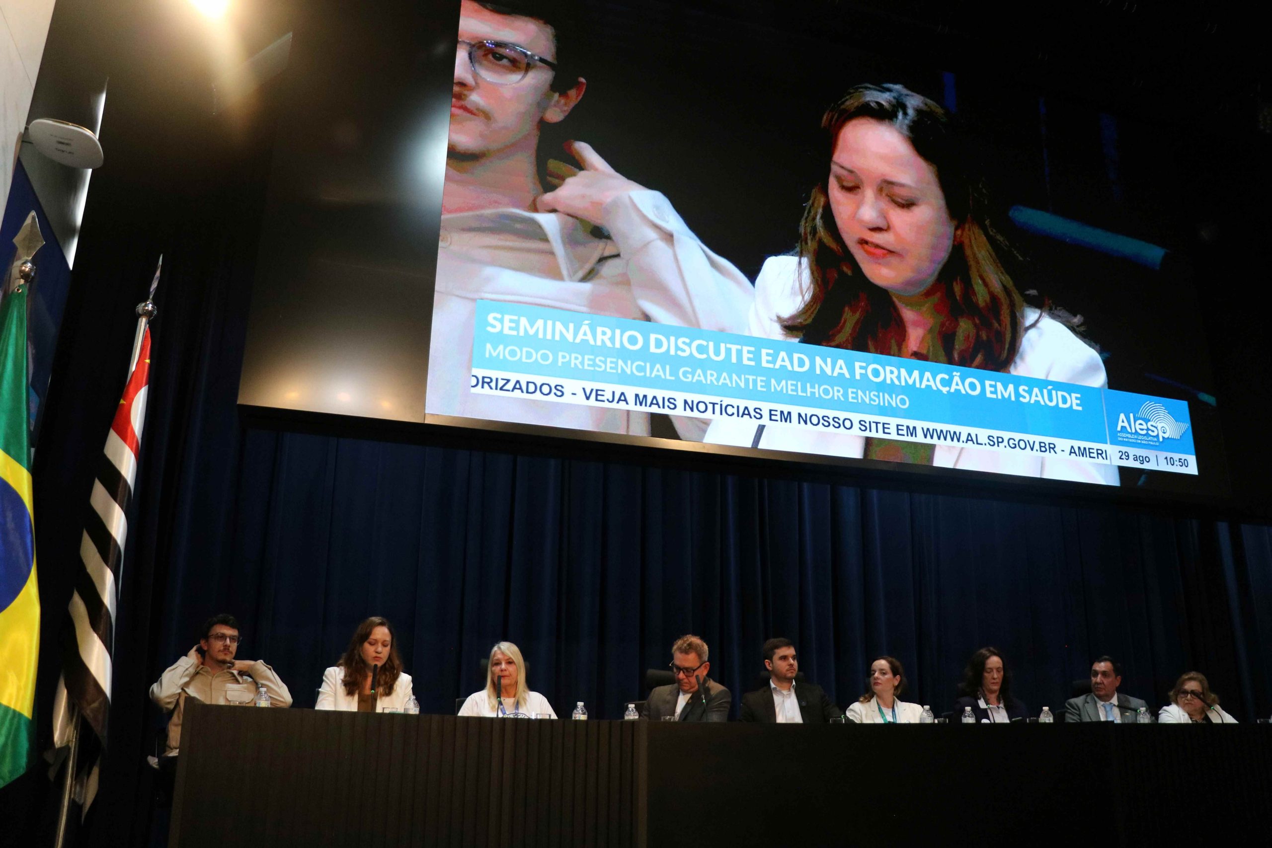 A presidente do CRMV-SP, Daniela Pontes Chiebao, sentada na mesa principal do seminário discursando. Ela aparece no telão que está acima da mesa principal. Ela veste blazer branco e blusa bege.
