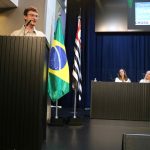 Homem branco jovem de fios castanhos lisos e curtos fala em um púlpito. Ele usa óculos de armação redonda e veste casaco bege. É o deputado estadual Guilherme Cortez.