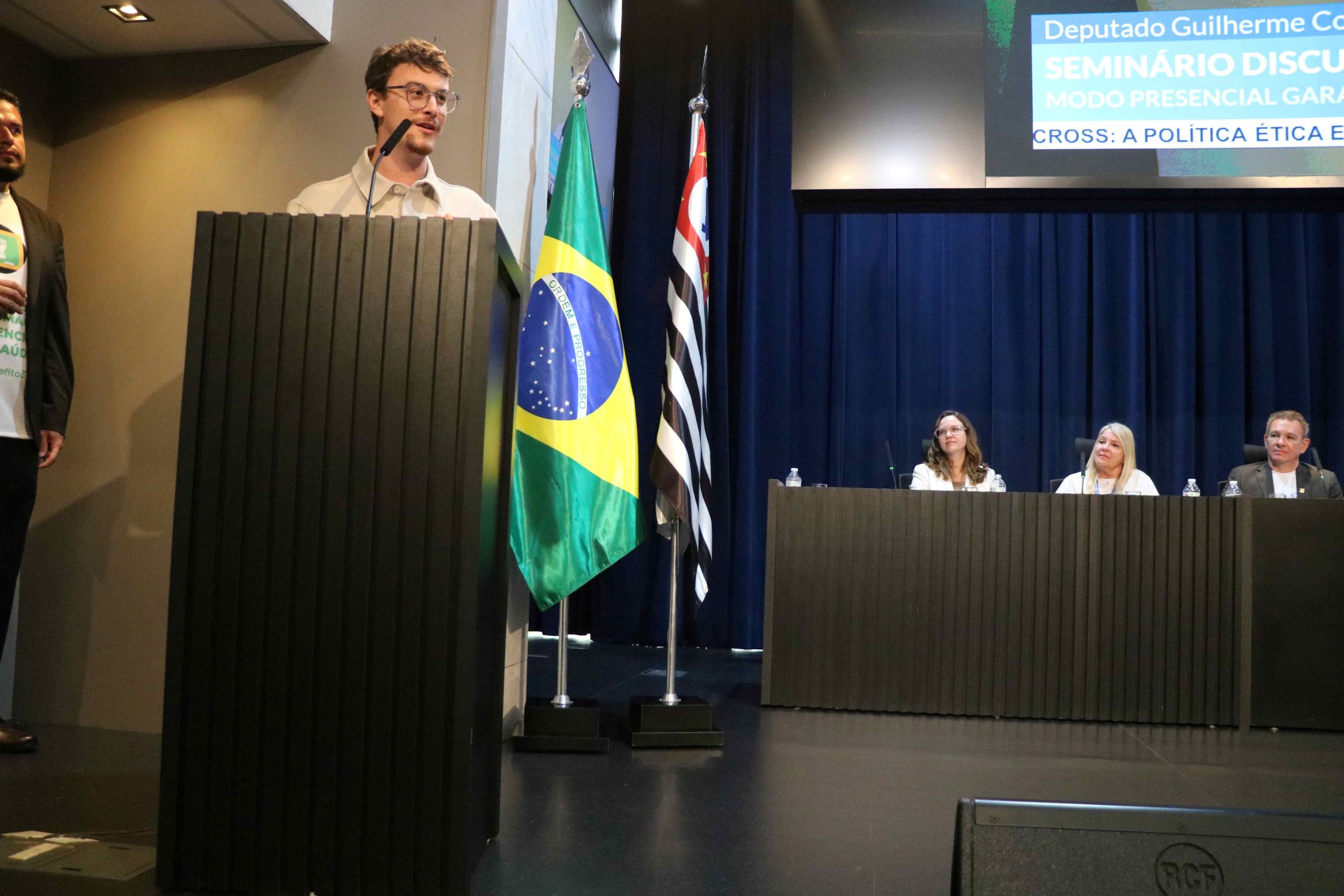 Homem branco jovem de fios castanhos lisos e curtos fala em um púlpito. Ele usa óculos de armação redonda e veste casaco bege. É o deputado estadual Guilherme Cortez.