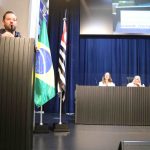 Homem branco de estatura mediana, fios pretos lisos e curtos fala no púlpito. Ele veste casaco preto. É o vereador Marcelo Messias.