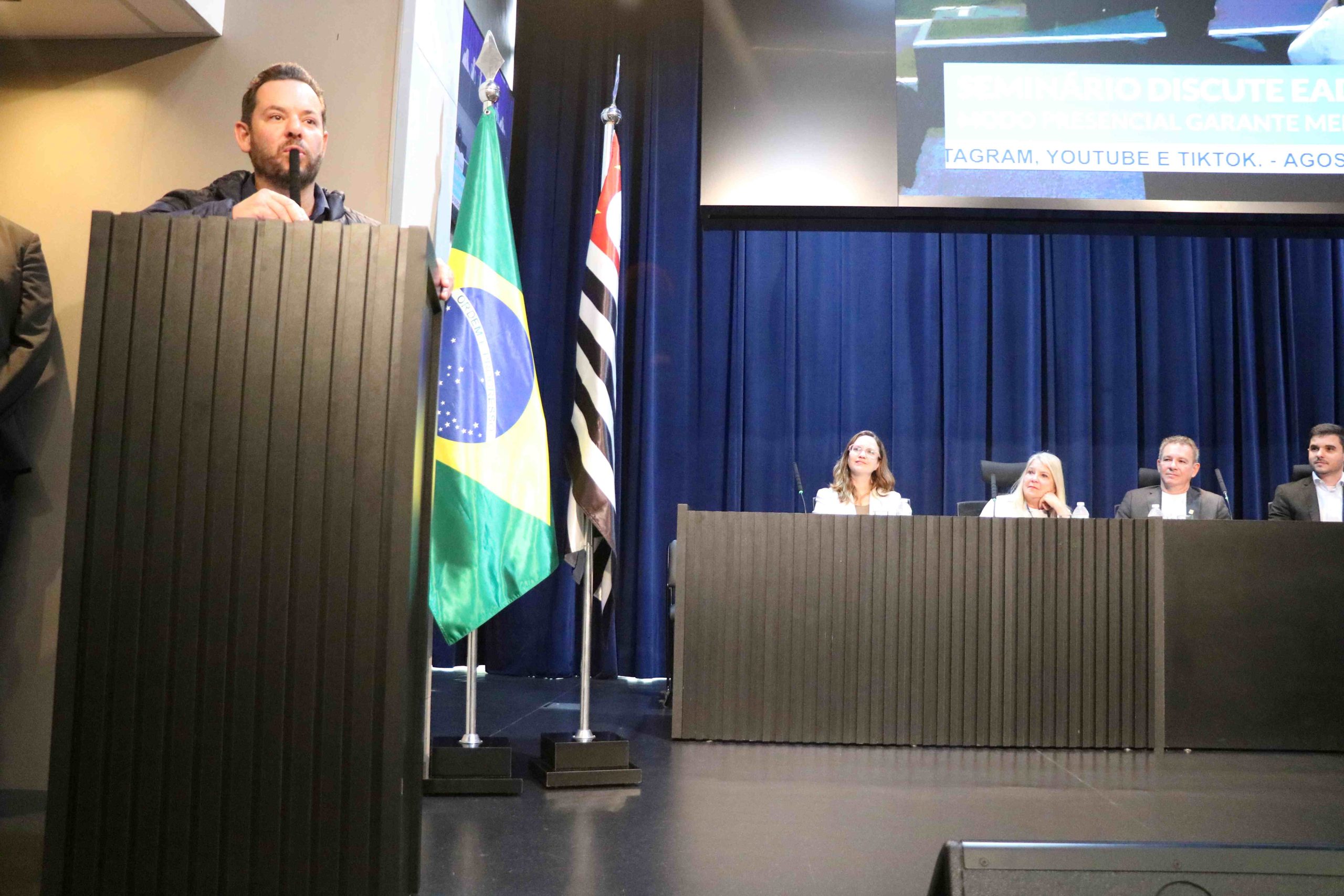 Homem branco de estatura mediana, fios pretos lisos e curtos fala no púlpito. Ele veste casaco preto. É o vereador Marcelo Messias.
