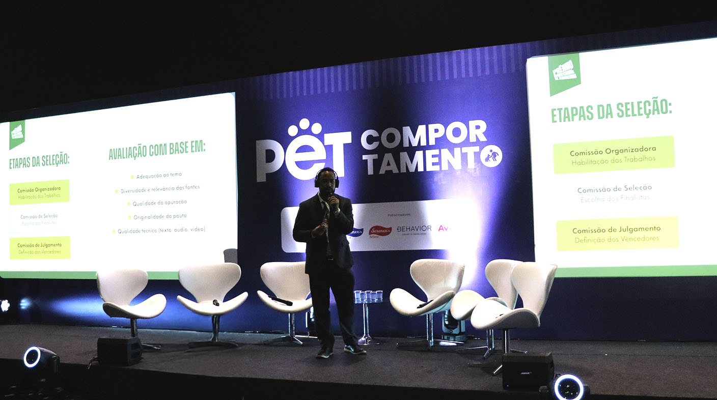 No palco do evento, Lucas Figueiredo, gerente de Comunicação do CFMV, de terno e tênis, fala ao microfone, durante o lançamento do Prêmio CFMV/CRMVs de Jornalismo.