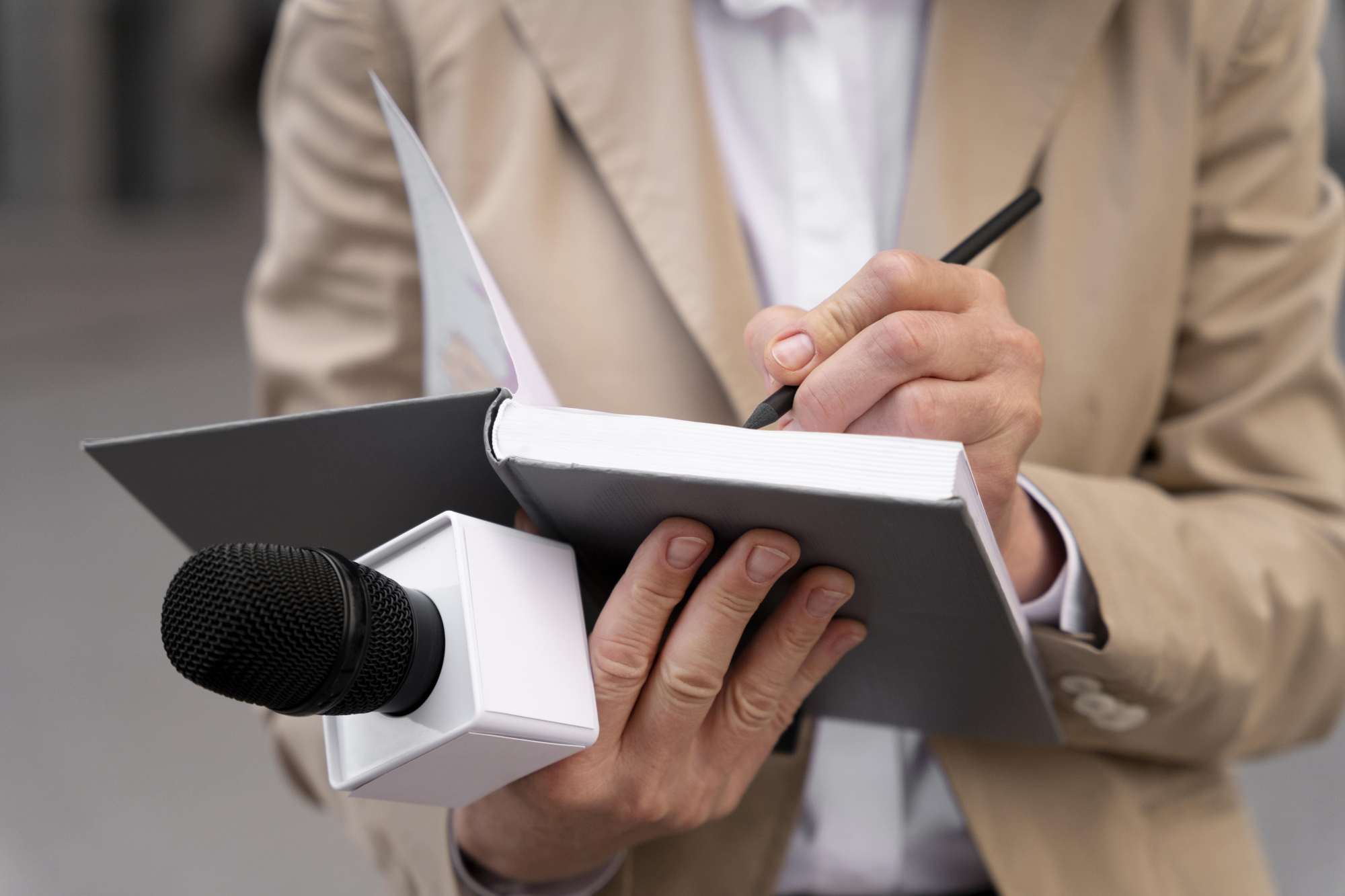 Corpo do pescoço para baixo, a pessoa, que aparenta ser um homem, veste um blazer creme e uma blusa branca de botões no meio. A imagem foca nas mãos brancas seguram uma agenda cinza aberta, um microfone e um lápis na mão direita.