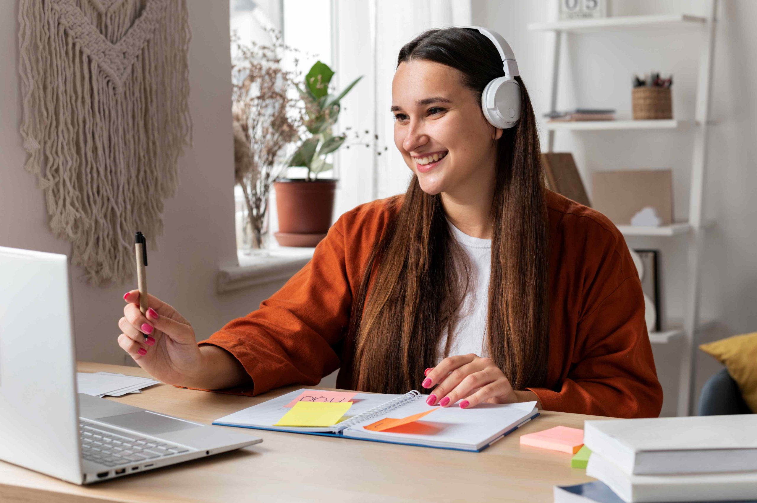 Mulher sorrindo em frente a um notebook, e na mesa alguns cadernos de anotação.
