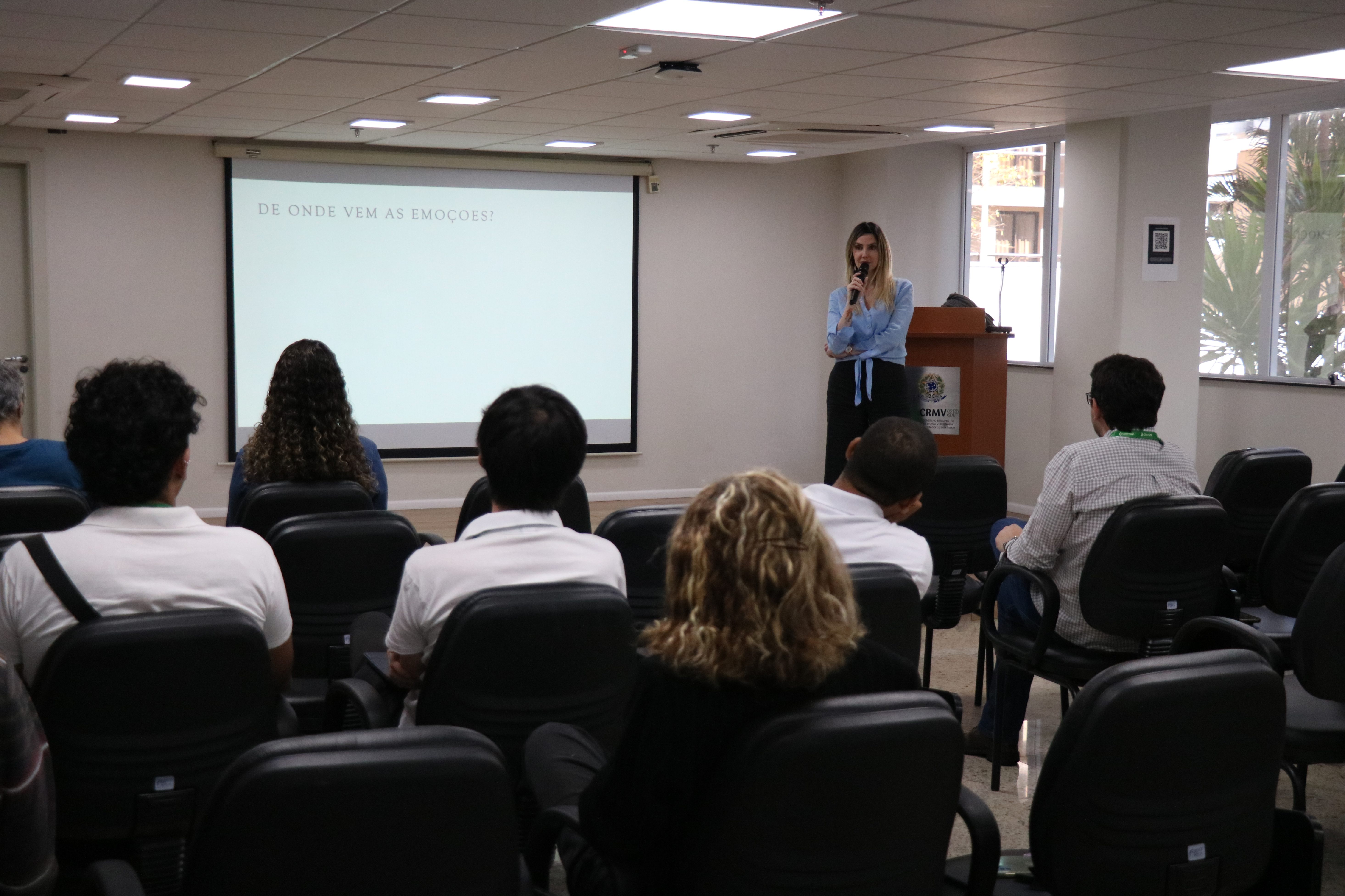 Funcionários do CRMV-SP sentados no auditório enquanto mulher branca de fios loiros lisos médios, vestindo blusa azul clara e calça preta social, fala no microfone. É a dra. Ingrid Bueno Atayde Machado.
